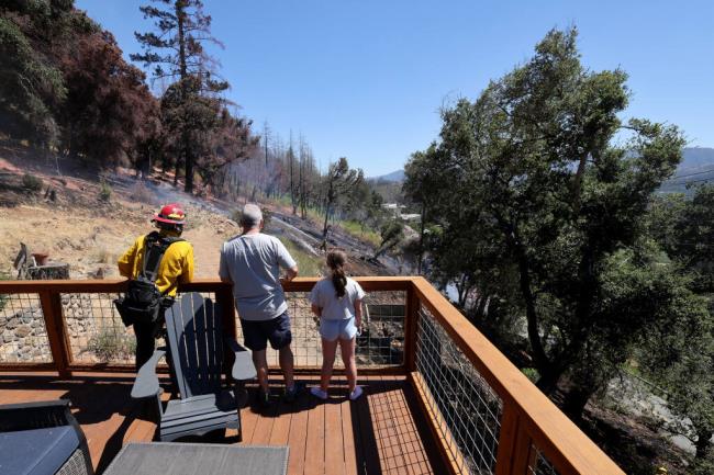 Jude Wilmoth, center, and his daughter Elyse, 9, watch from the deck of their home as firefighters from Cal Fire and the Deer Park Volunteer Fire Department work to put out a vegetation fire off Sanitarium Road in Deer Park, Sunday, July 30, 2023. The fire came within a few feet of their house. (Beth Schlanker / The Press Democrat)